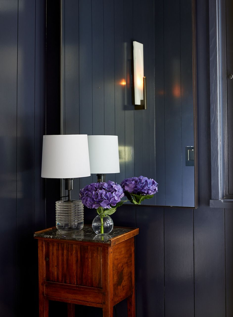 Un miroir reflète une applique murale contemporaine, une petite lampe avec un abat-jour rond blanc et un vase en verre avec des fleurs bleues, qui repose sur une table d’appoint.
