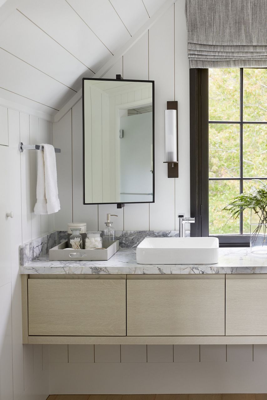 Détail de la baignoire à Ray Booth, avec un miroir à cadre noir accroché à un angle, au-dessus du dessus de meuble-lavabo en marbre avec lavabo en surface Vox rectangulaire, accessoires de baignoire sur un plateau et un vase avec de la verdure.