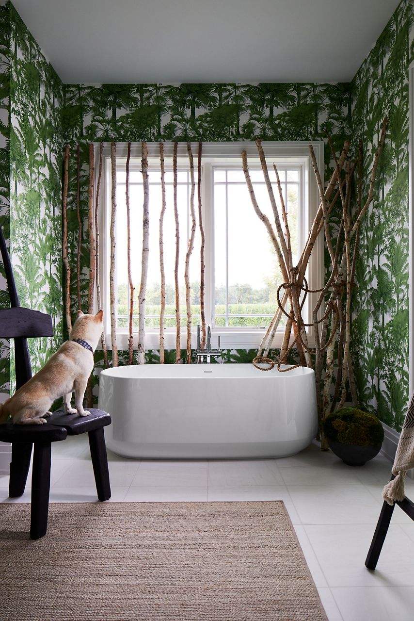 A dog sits on a chair in a tropical bathroom and looks at a KOHLER Ceric freestanding tub