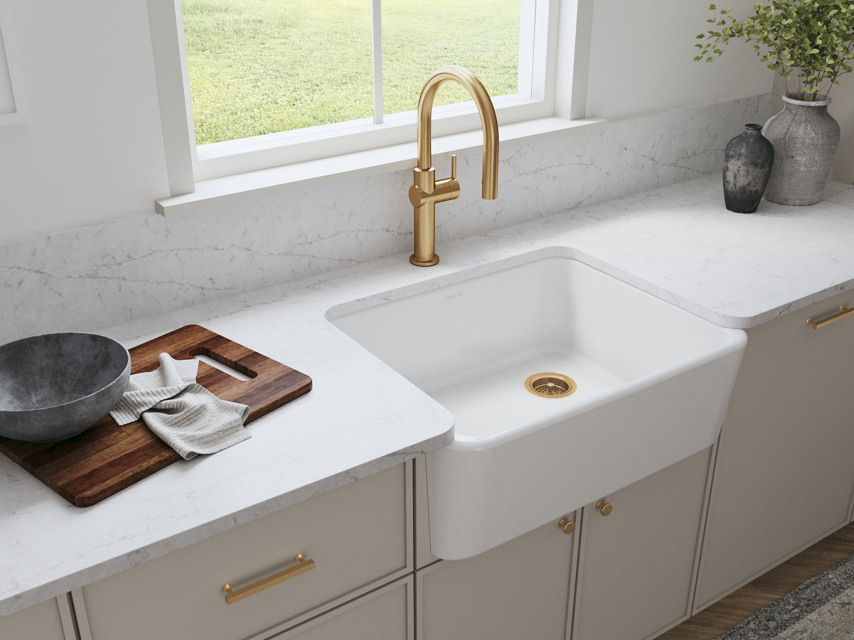 A kitchen with white counters, light cabinets, and a white KOHLER farmhouse sink installed underneath a large window.