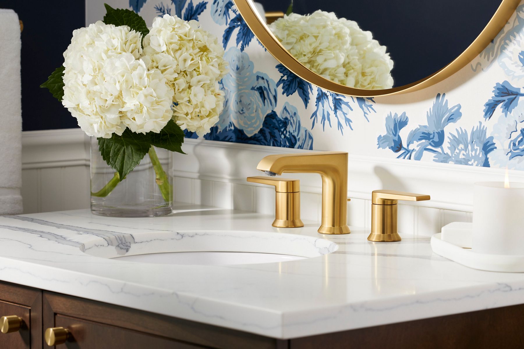A brushed brass bathroom faucet on a white counter top with blue floral wallpaper behind it.