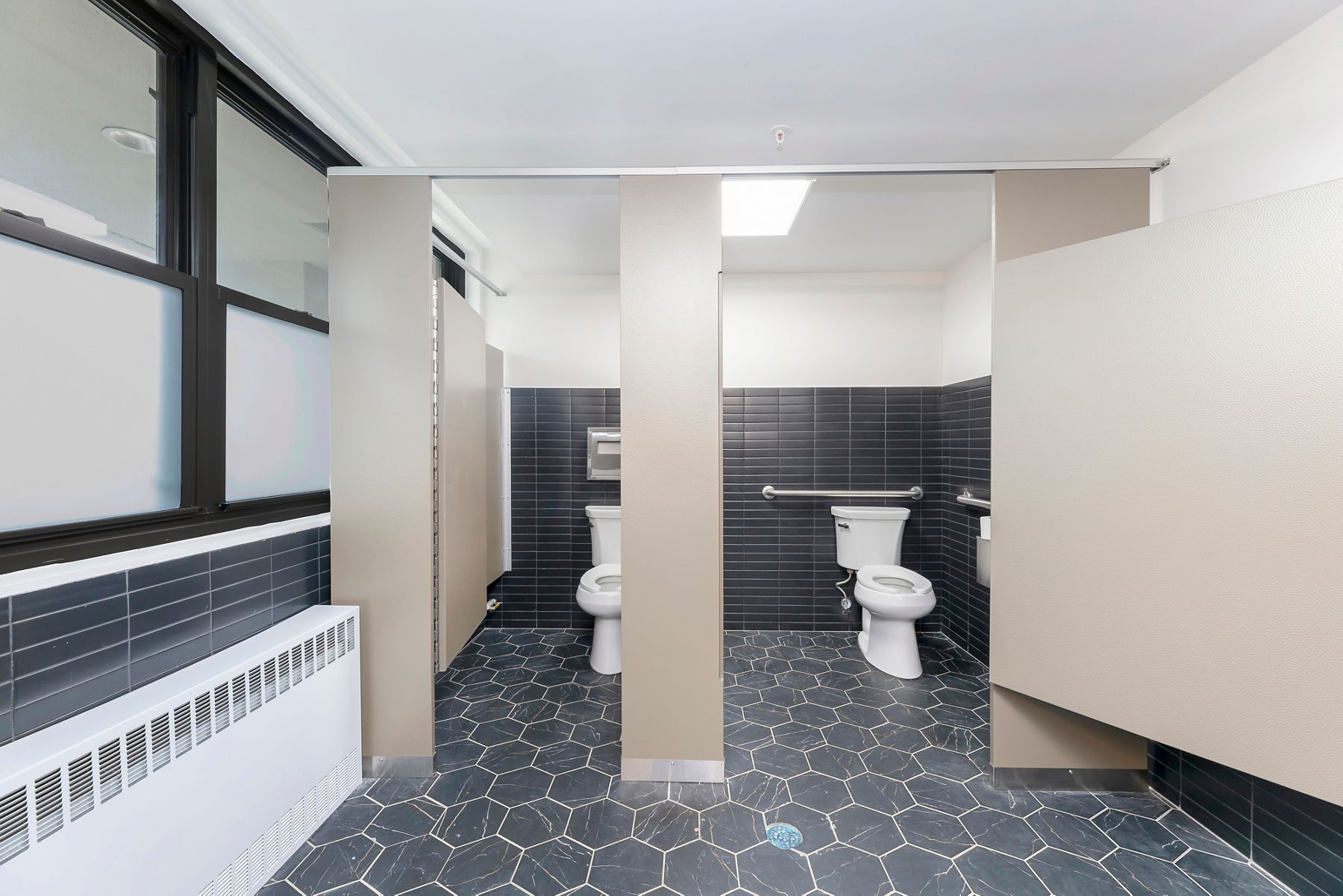 Updated shared restroom with modern dark floor tile, white partitions, and multiple stalls.