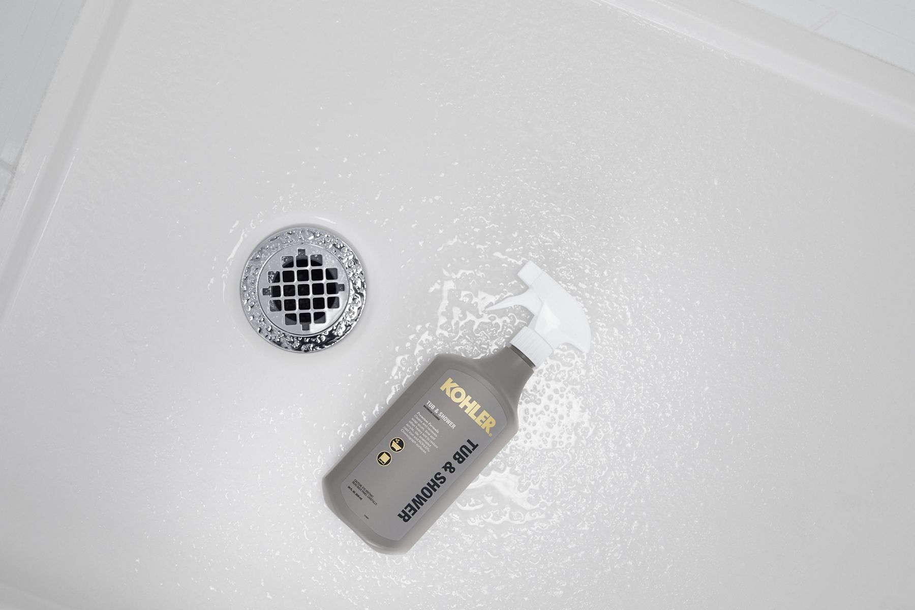 A spray bottle of KOHLER Tub and Shower Cleaner lies on its side next to the drain of an acrylic shower base