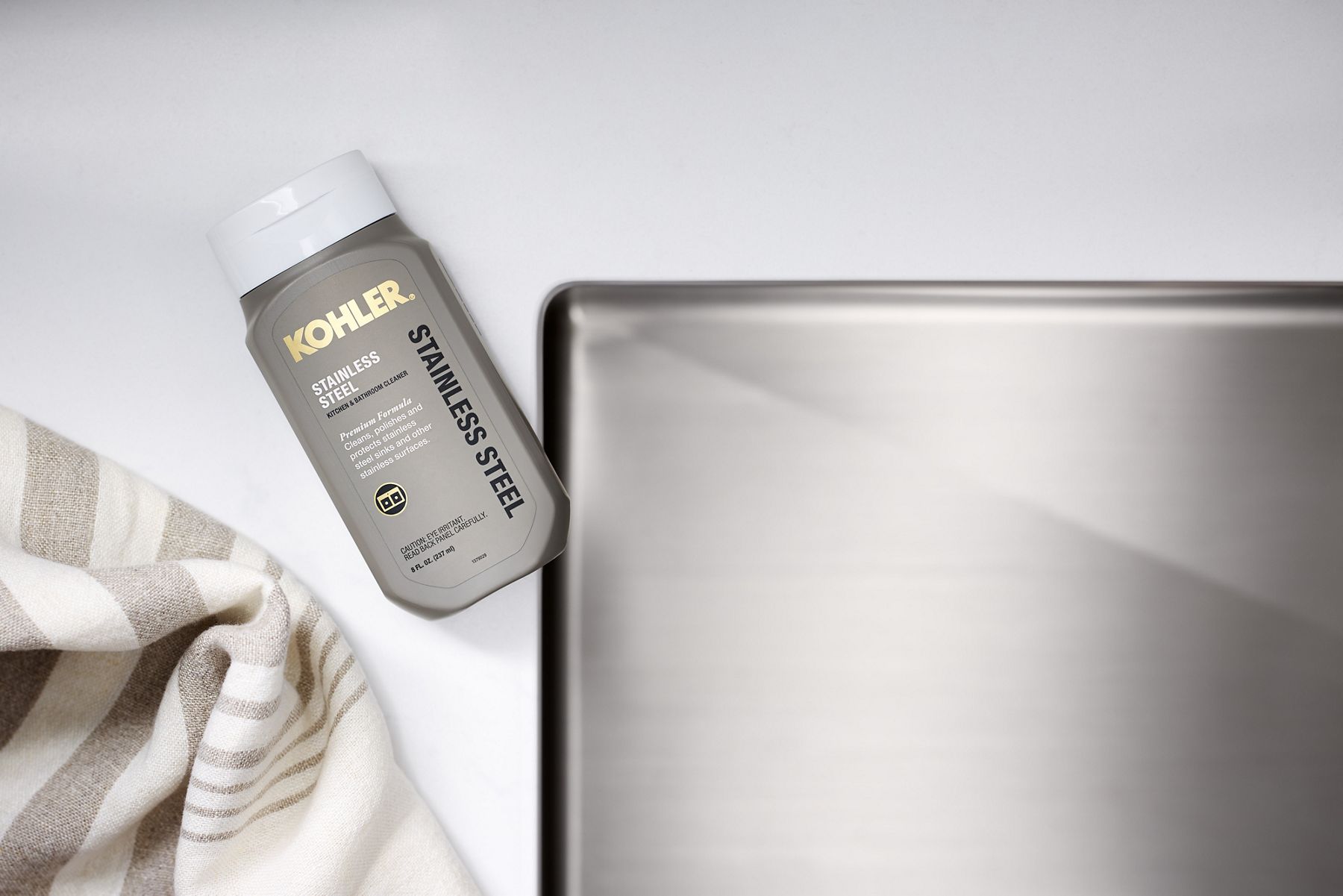 A bottle of KOHLER stainless steel cleaner with corner of a stainless steel sink and white and gray tea towel.