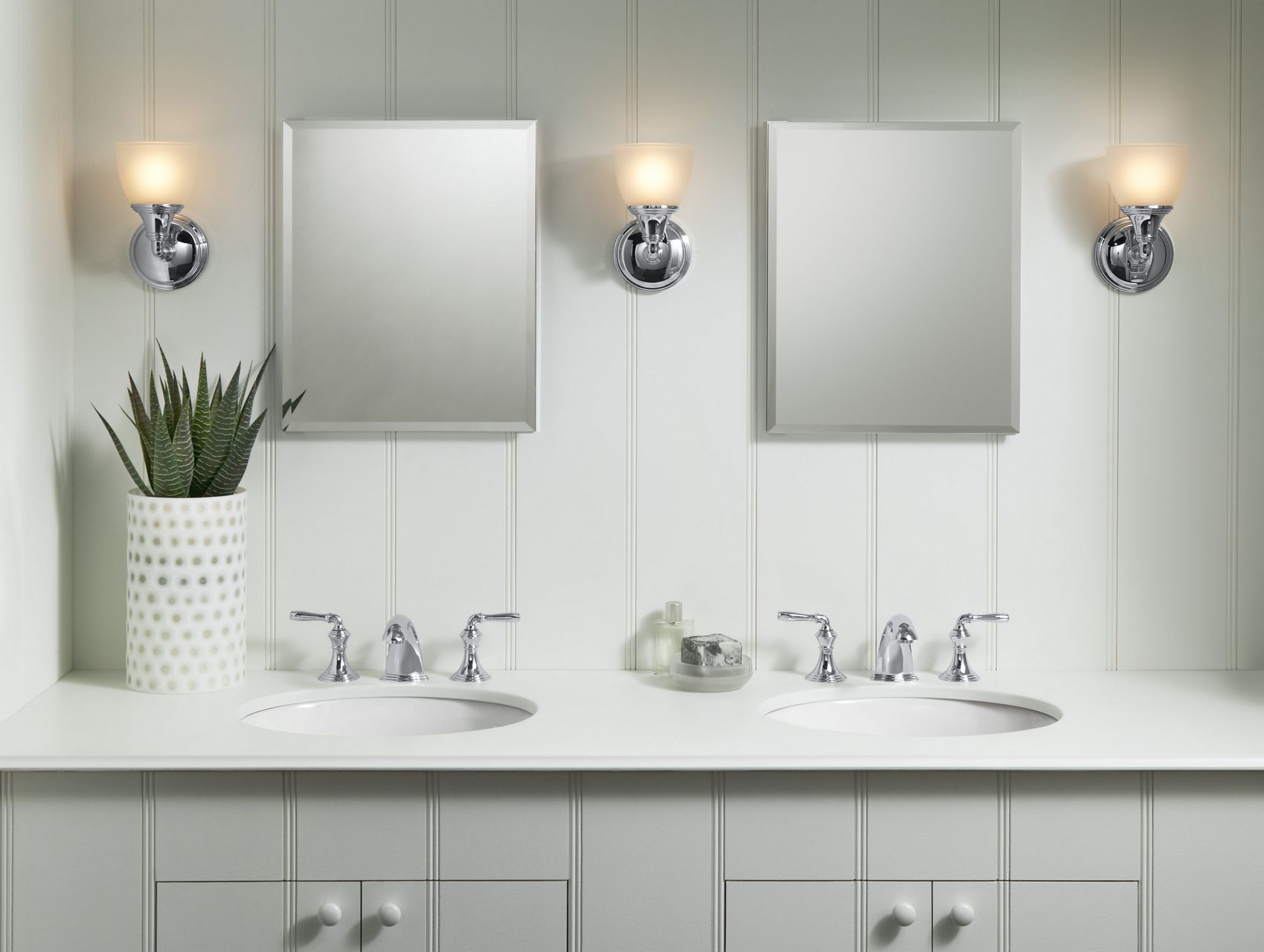 A light gray double vanity against a light gray wall with a white counter top, chrome faucets, mirrors and lighting.