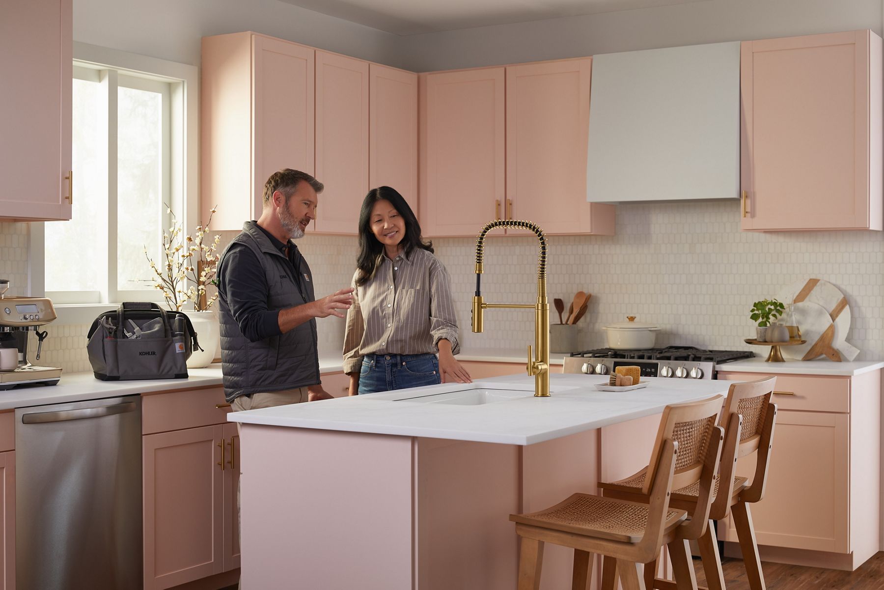 A workman and a woman in a pink and white kitchen looking at and discussing a brushed brass faucet he just installed.