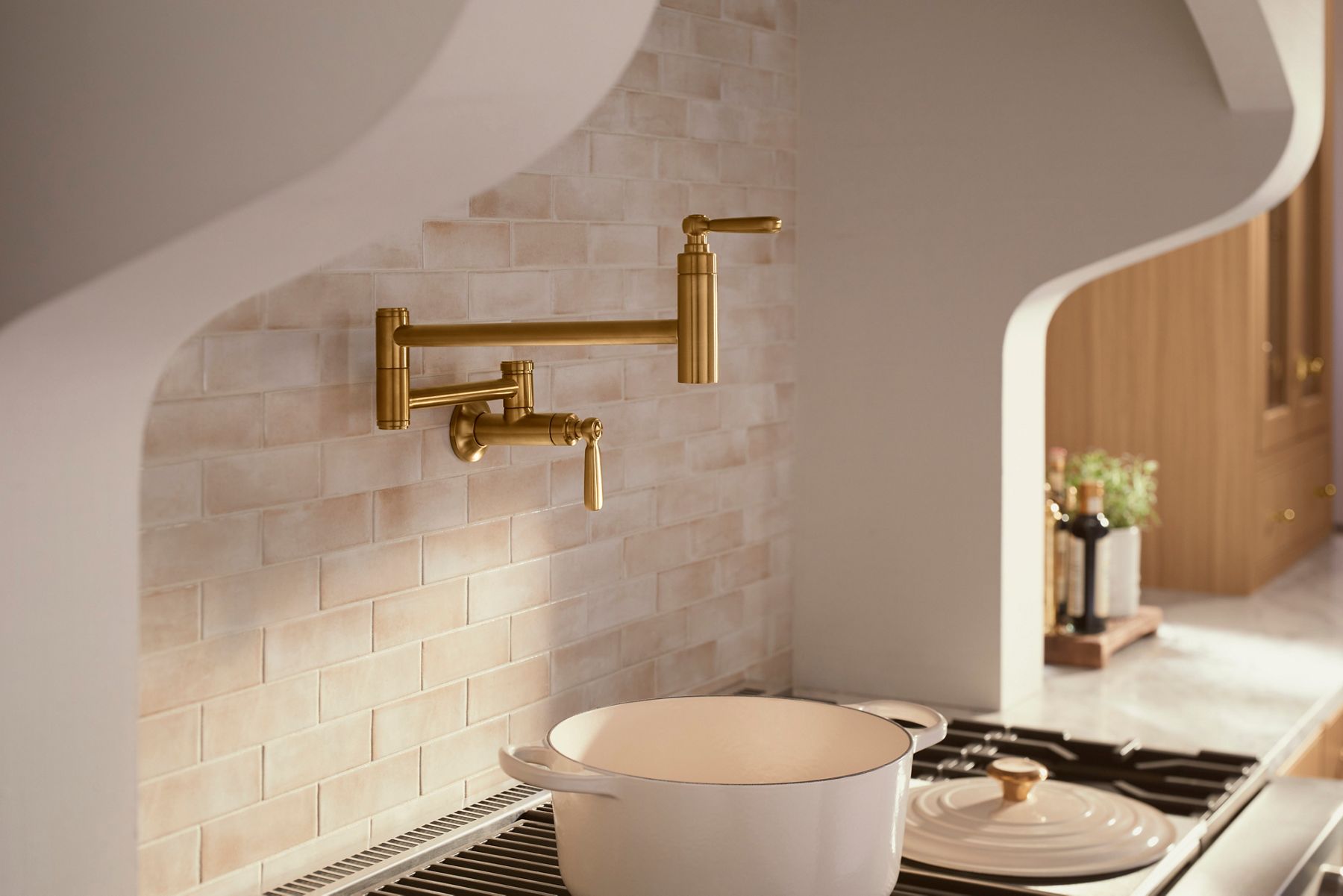A luxurious kitchen featuring a gold wall-mounted pot filler faucet above a stove with a large cream-colored Dutch oven. The backsplash consists of light beige subway tiles, and the space is designed with a soft, warm color palette. The arched hood adds an elegant touch, framing the cooking area beautifully.