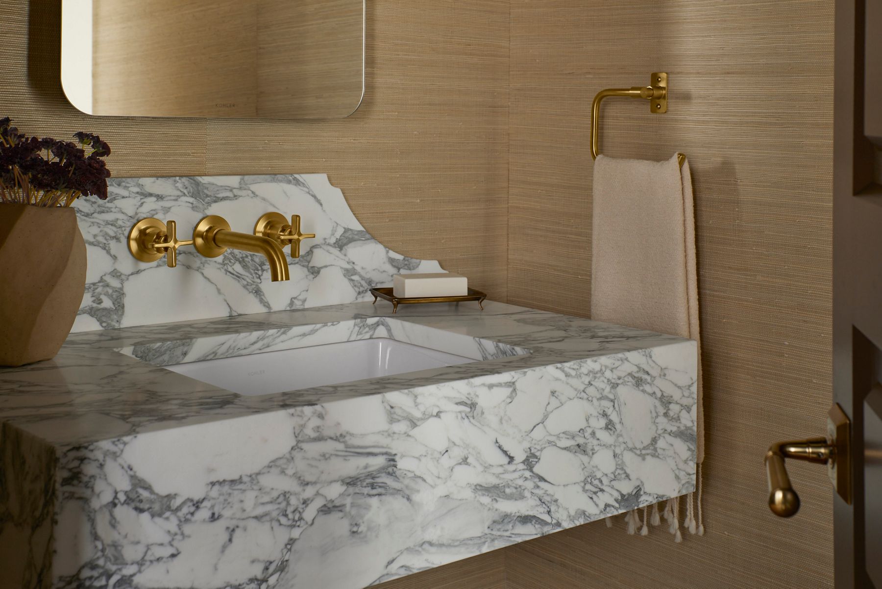 A close-up of a wall-mounted brass bathroom faucet with cross handles and curved spout installed above a veined marble back-splash and integrated sink