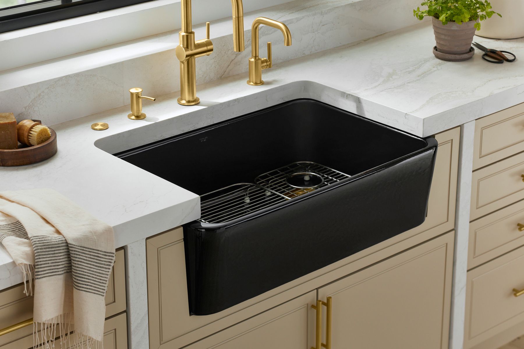A modern kitchen featuring a black farmhouse-style sink with a white marble countertop. The sink area includes a set of gold faucets, including a high-arc spout, a smaller faucet, and a soap dispenser, all matching in design. The cabinetry below is a light beige color, adding a touch of warmth to the space, while the overall design is elegant and luxurious.
