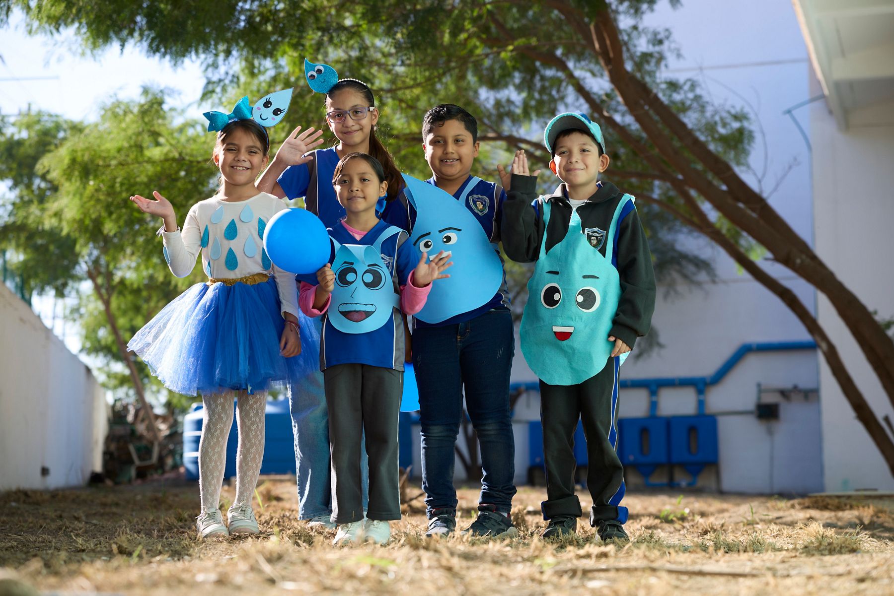 Five children outdoors wearing blue water droplet-themed costumes and accessories, smiling and posing together with blue balloons and cartoon droplet cutouts.