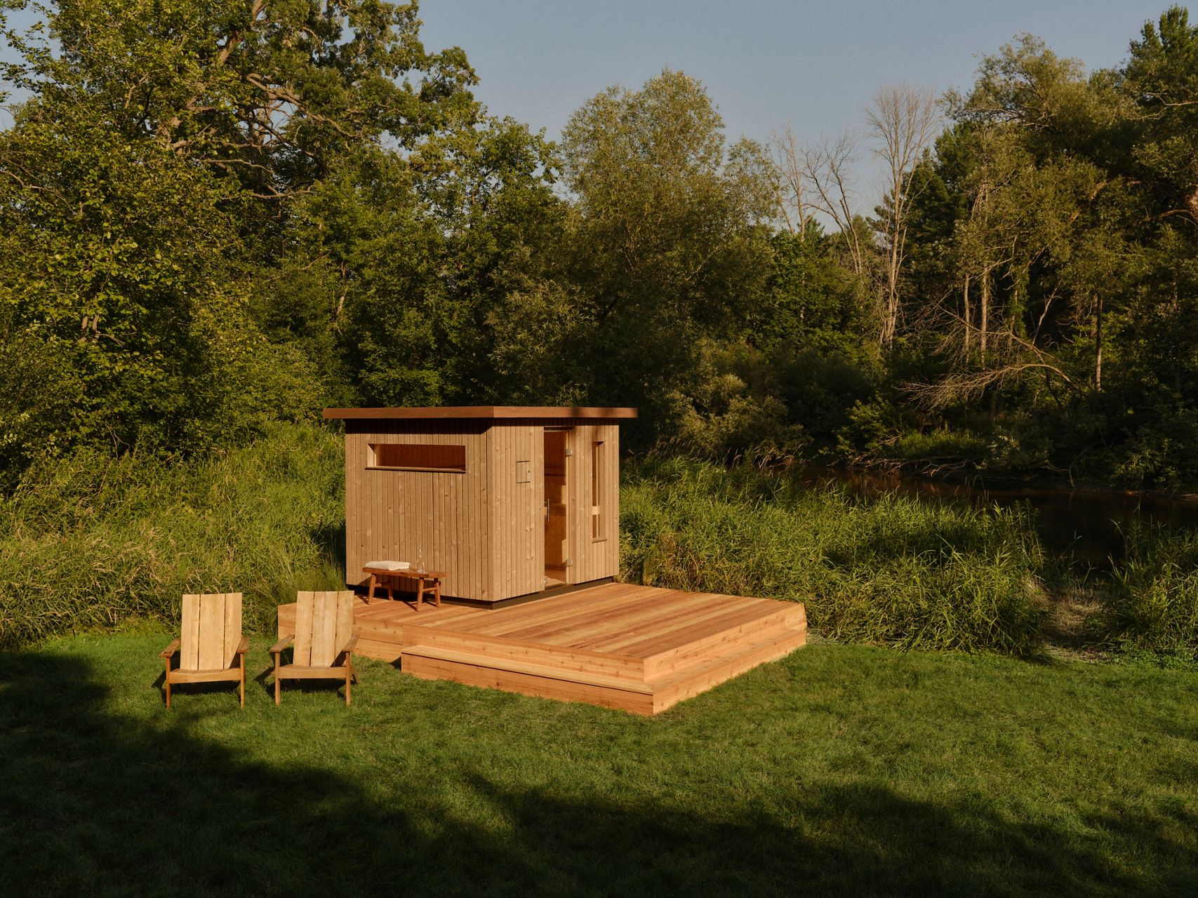 Wooden sauna cabin with deck and chairs, surrounded by lush greenery.