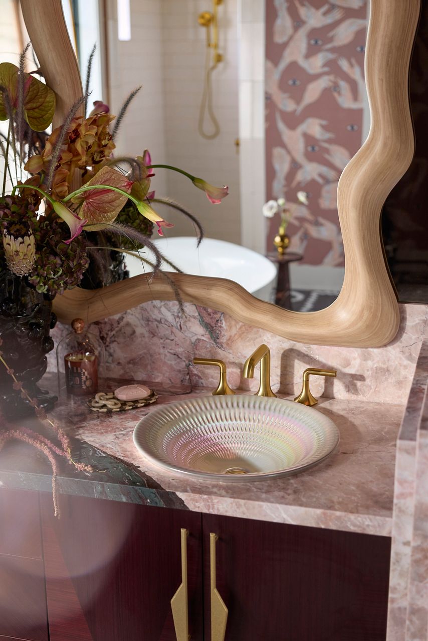 Multicolored ceramic and glass sink with a pink marble counter, gold fixtures, and a curved mirror.