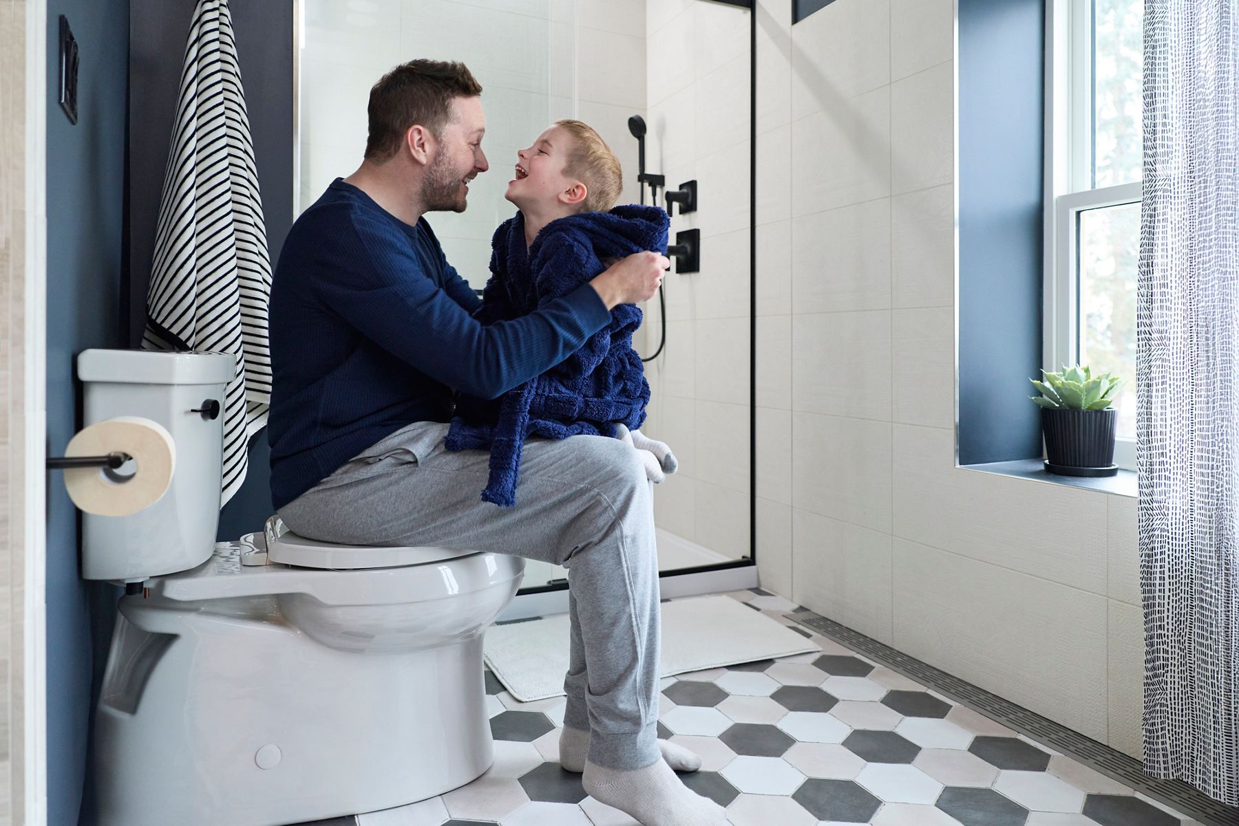 Adult and child sharing a playful moment in a bright modern bathroom, seated on a white toilet beside a glass shower enclosure with hexagon tile flooring and a window ledge with a small plant.