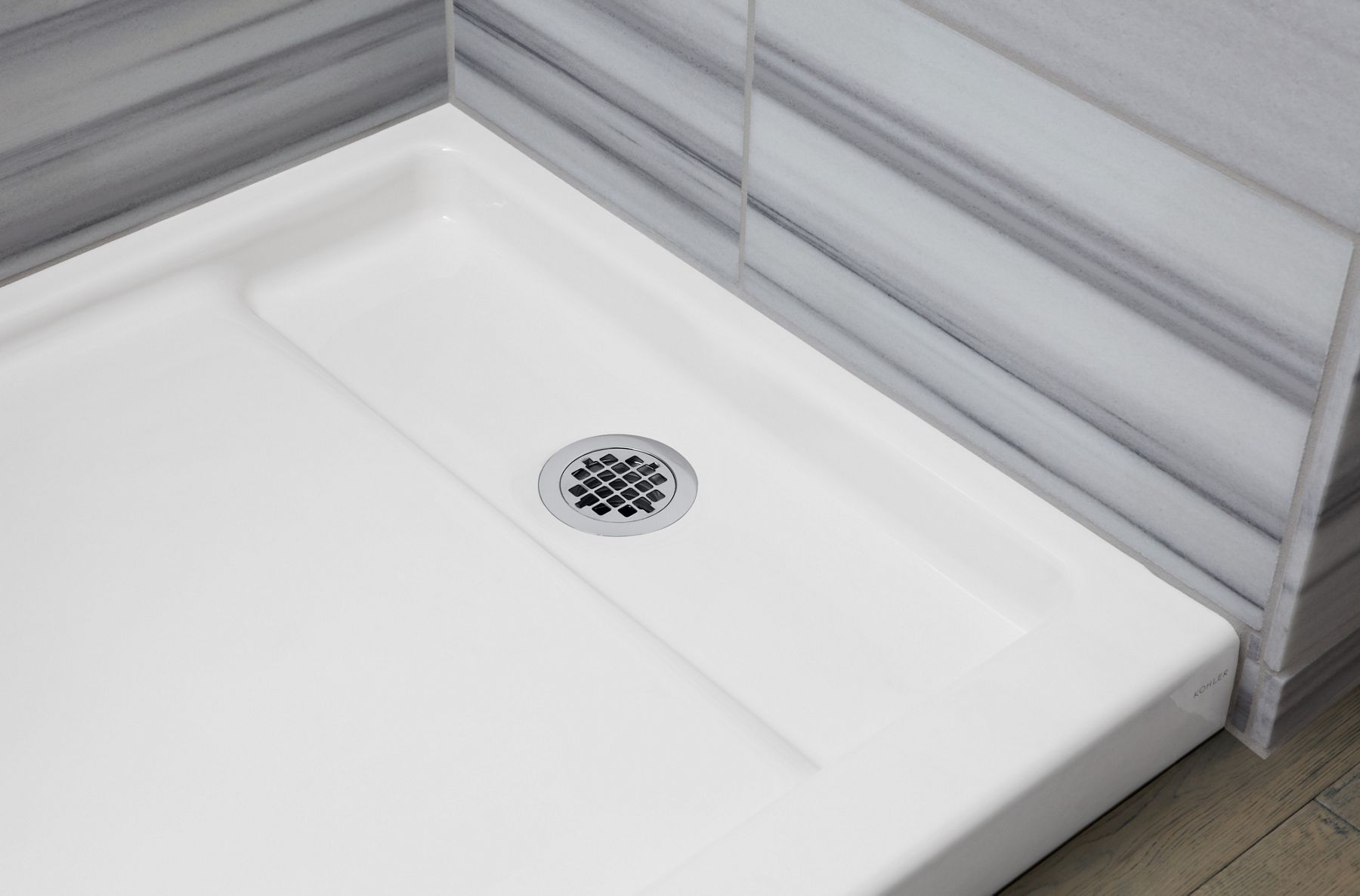 A close-up of a white enameled cast iron shower base and drain backed a gray wall