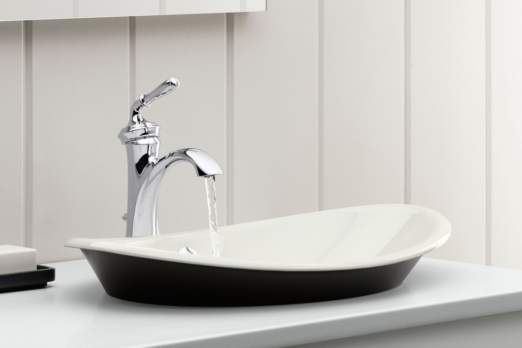 A close-up of a chrome bathroom sink faucet with running water over an enameled cast iron bathoom sink with white bowl and black base