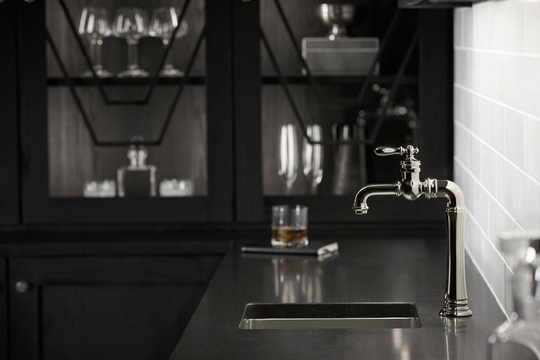 A sophisticated kitchen or bar area featuring a vintage-inspired polished chrome faucet with intricate detailing. The faucet is mounted on a dark countertop with an undermount sink. In the background, there is a glass of whiskey and a dark cabinet with glassware, creating a moody and elegant atmosphere. The white subway tile backsplash contrasts with the dark tones, adding a touch of brightness to the space.