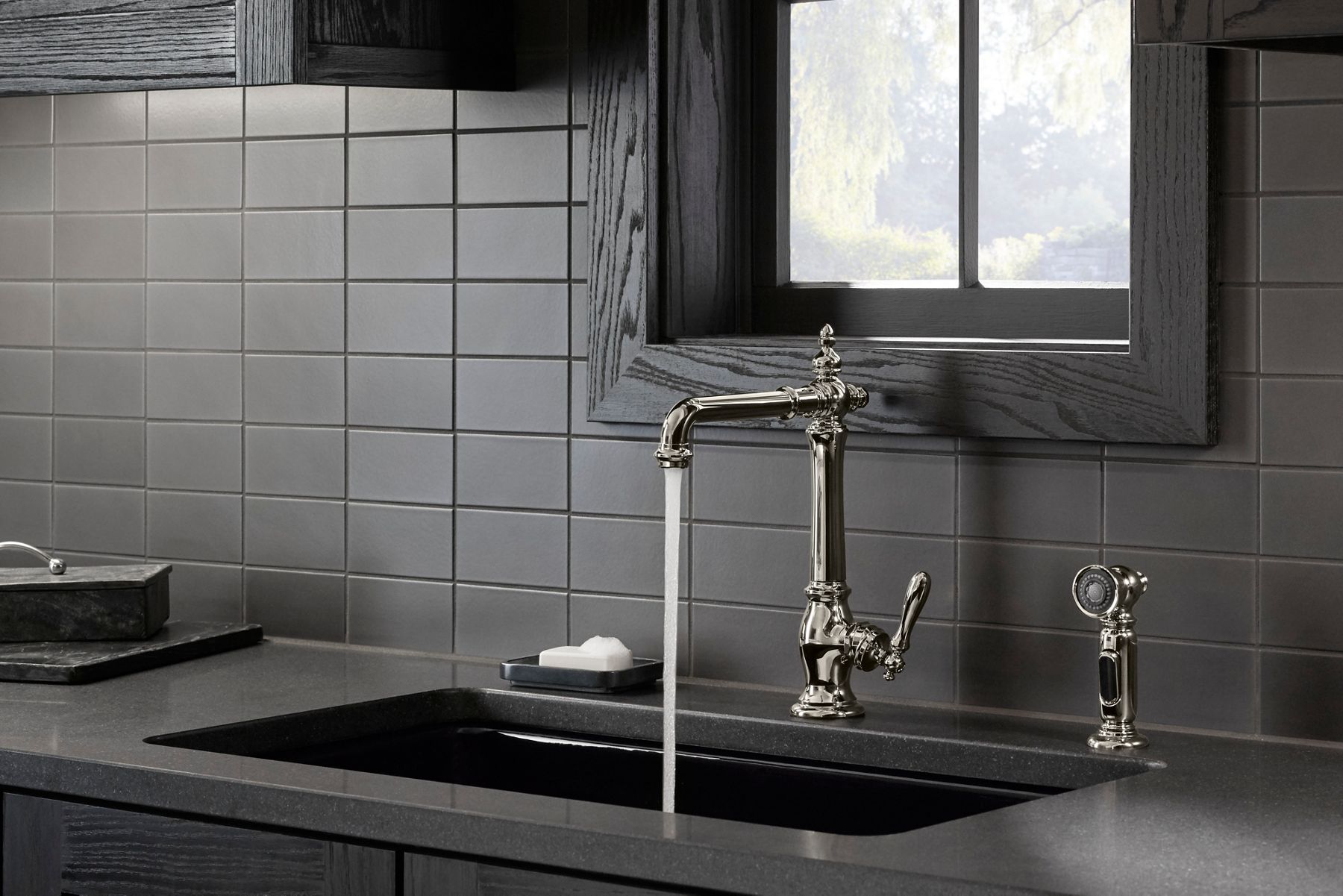 Traditional kitchen with a vintage-inspired polished chrome faucet featuring intricate detailing and a matching side spray. Water flows from the faucet into a dark countertop sink. The backsplash consists of dark grey subway tiles, and a soap dish with a white bar of soap is placed nearby, enhancing the classic and elegant style of the space.