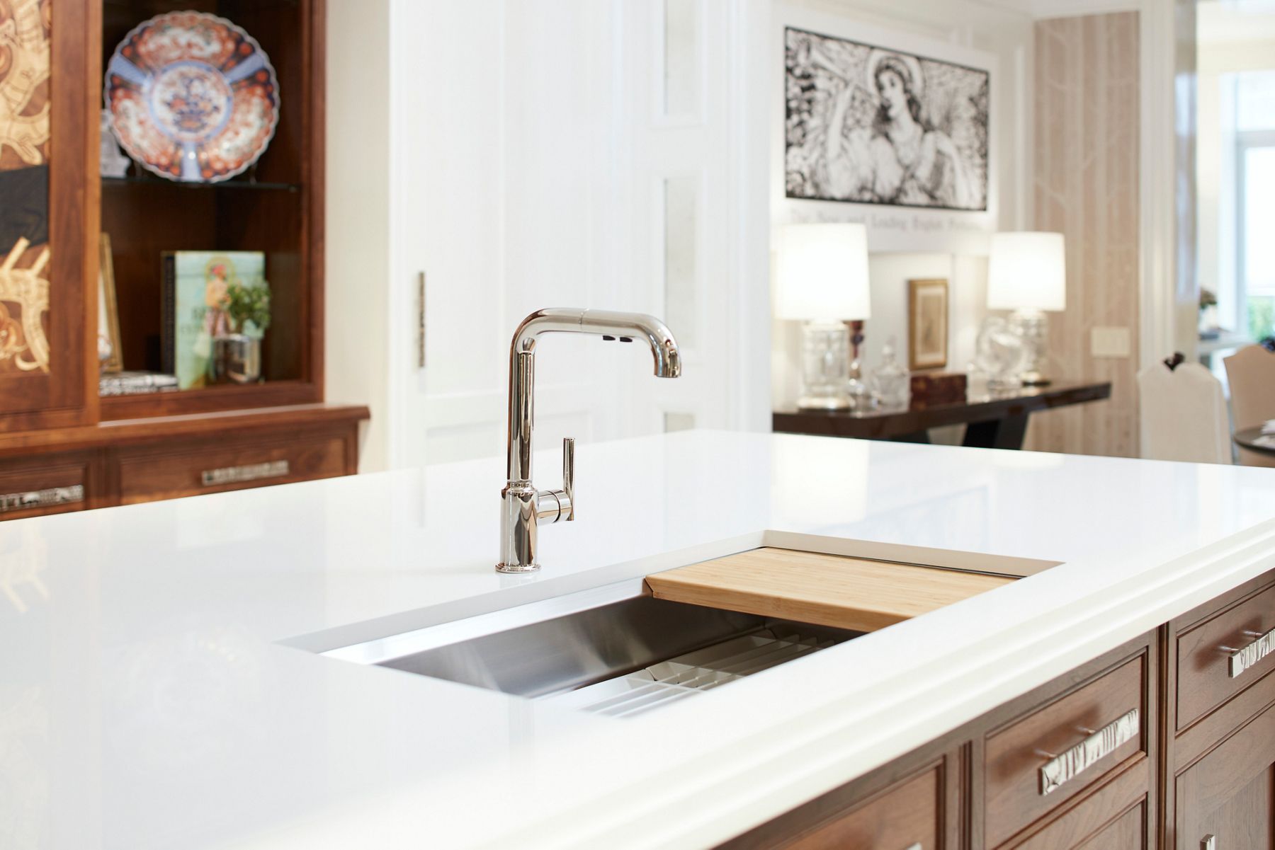 Elegant kitchen island with a polished chrome faucet and an integrated stainless steel sink. The white countertop is complemented by a wooden cutting board placed over the sink. In the background, decorative elements like artwork, table lamps, and a display cabinet with fine china add a touch of sophistication to the space.