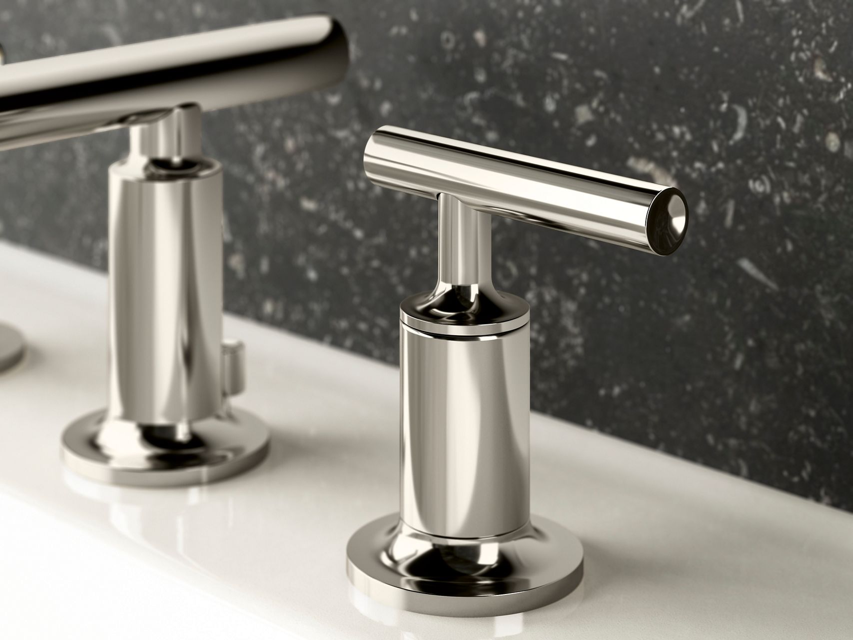 A close-up of chrome bathroom sink lever handles on a white sink, backed by black granite