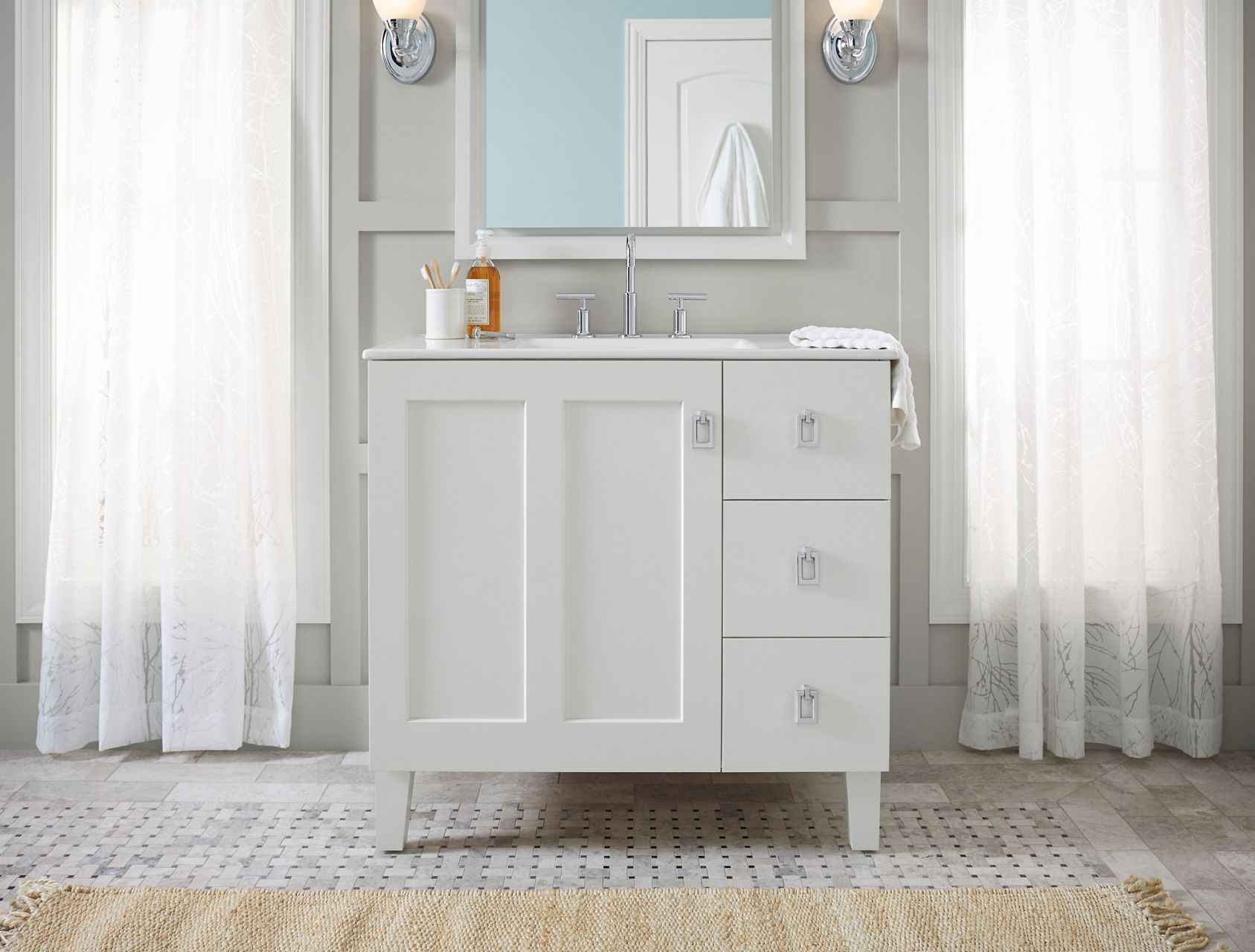 A white vanity with a large door on the left and a column of three drawers on the right, a rectangular mirror, sconce lights and floor-length lace curtains on either side.
