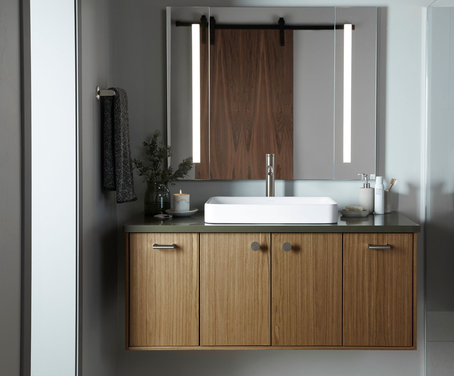 A bathroom styling space with a medium wood tone wall-mount vanity with four equal size doors, a green counter top, rectangular white vessel sink, chrome faucet, and a lighted mirror.