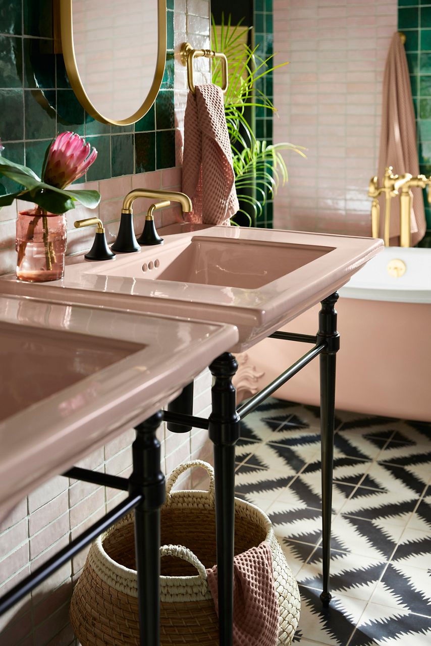 A double pale bathroom sink with black and gold faucets. There's a pale pink tub in the back with a gold faucet. The floors are patterned and the walls are tiled with emerald green.