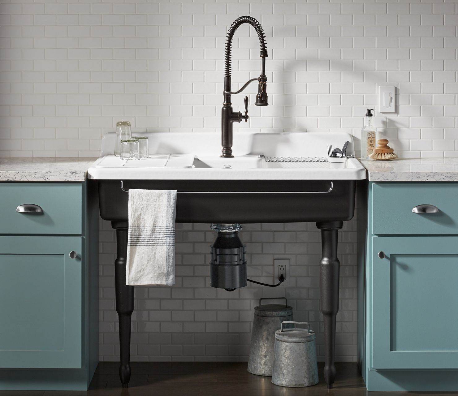 An enameled cast iron Farmstead sink with black exterior and legs and a white enameled interior, set in a pastel teal cabinet with white tile backsplash and a semiprofessional faucet.