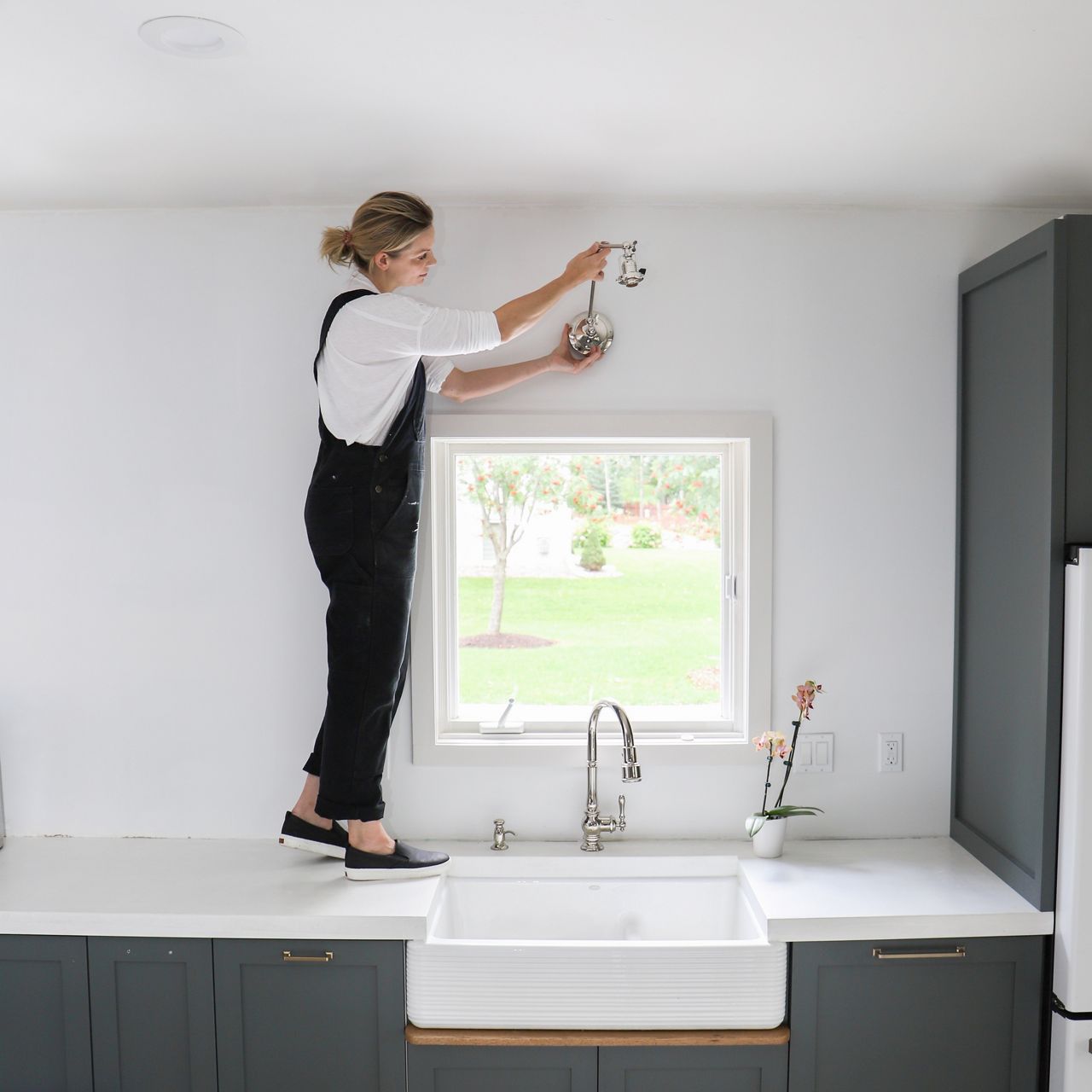 Jennifer Gizzi stands on a white kitchen countertop to install a single light sconce over a window and white farmhouse sink.
