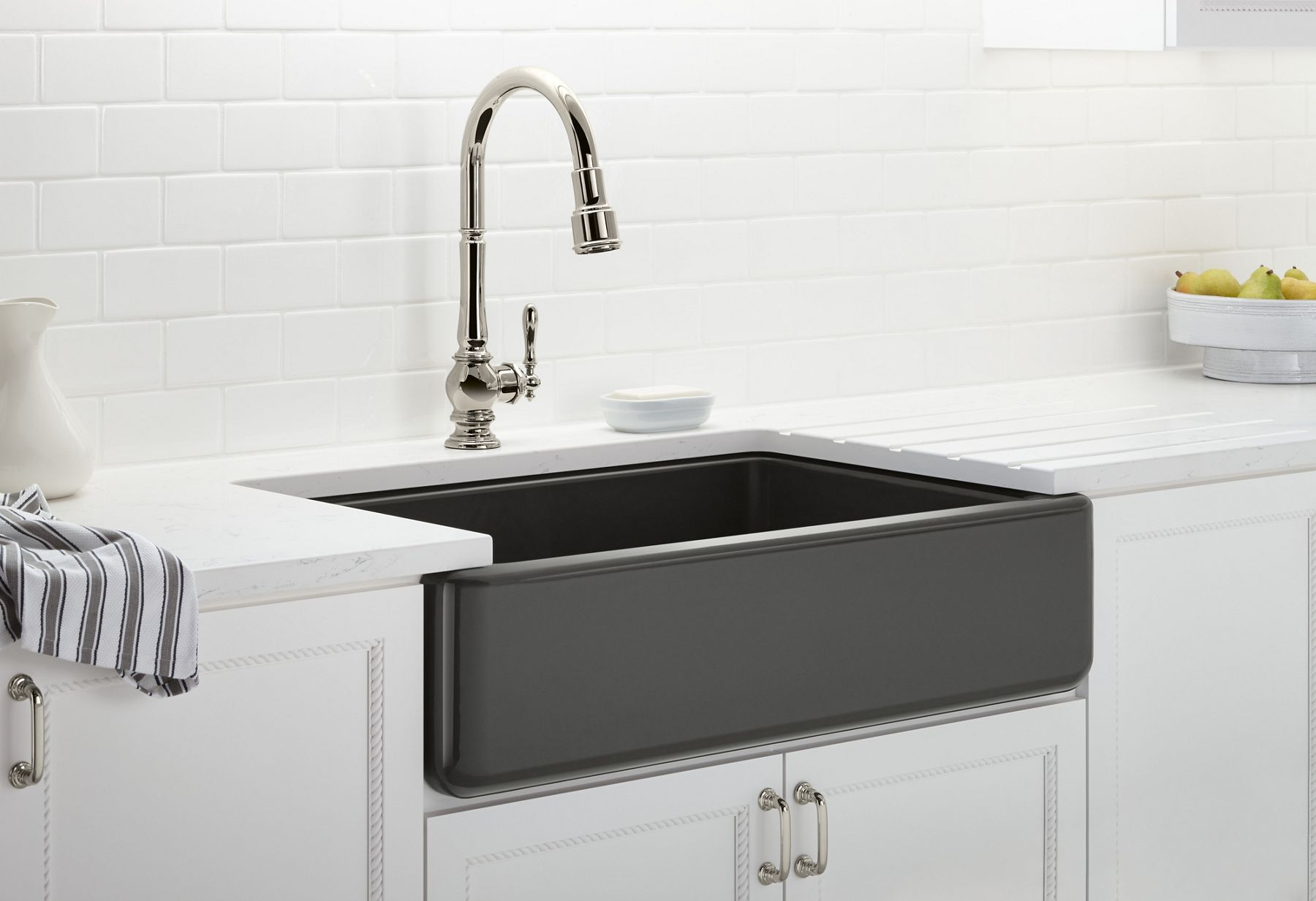 Delicate teacups are stacked next to a KOHLER tall-apron farmhouse stainless steel sink set in wooden cabinetry and marble countertops