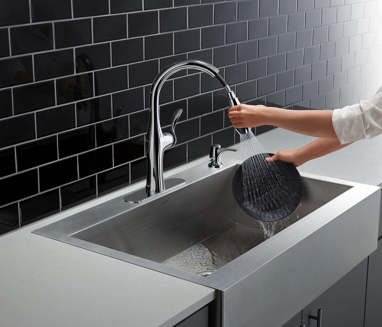 Hands wash a dirty pan in a KOHLER Vault stainless steel kitchen sink with shortened apron-front using a pull-down faucet in a high-contrast kitchen