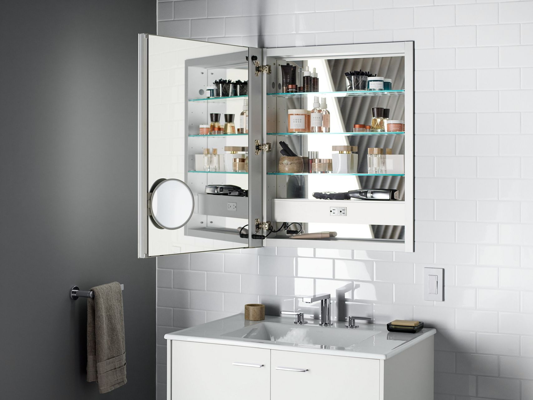 A medicine cabinet with its door open to show a mirrored inside door and back panel, along with a magnification mirror, an electrical outlet, and storage for numerous daily items, all set against a white tile wall and over a white vanity.