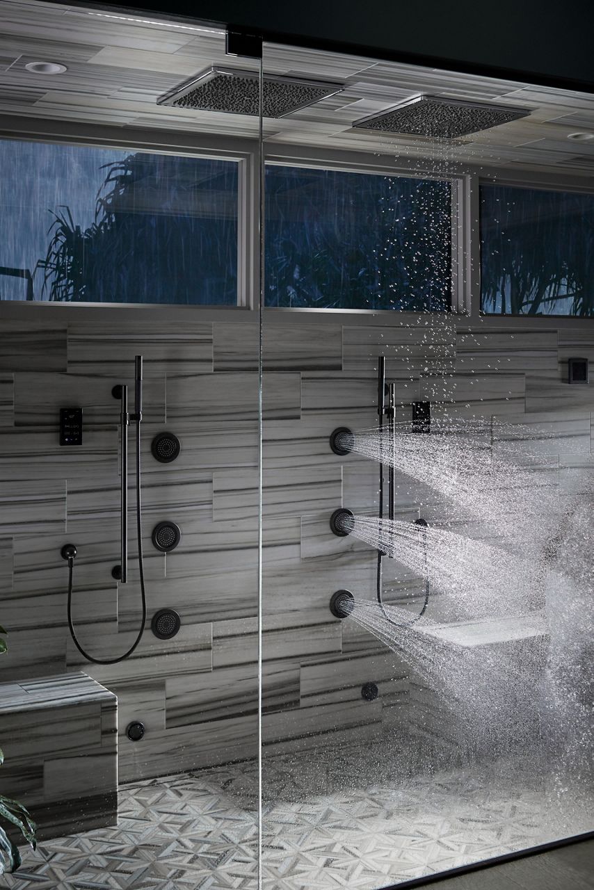 Water shooting out of three vertical body sprays in a shower area with horizontally striped gray tile.