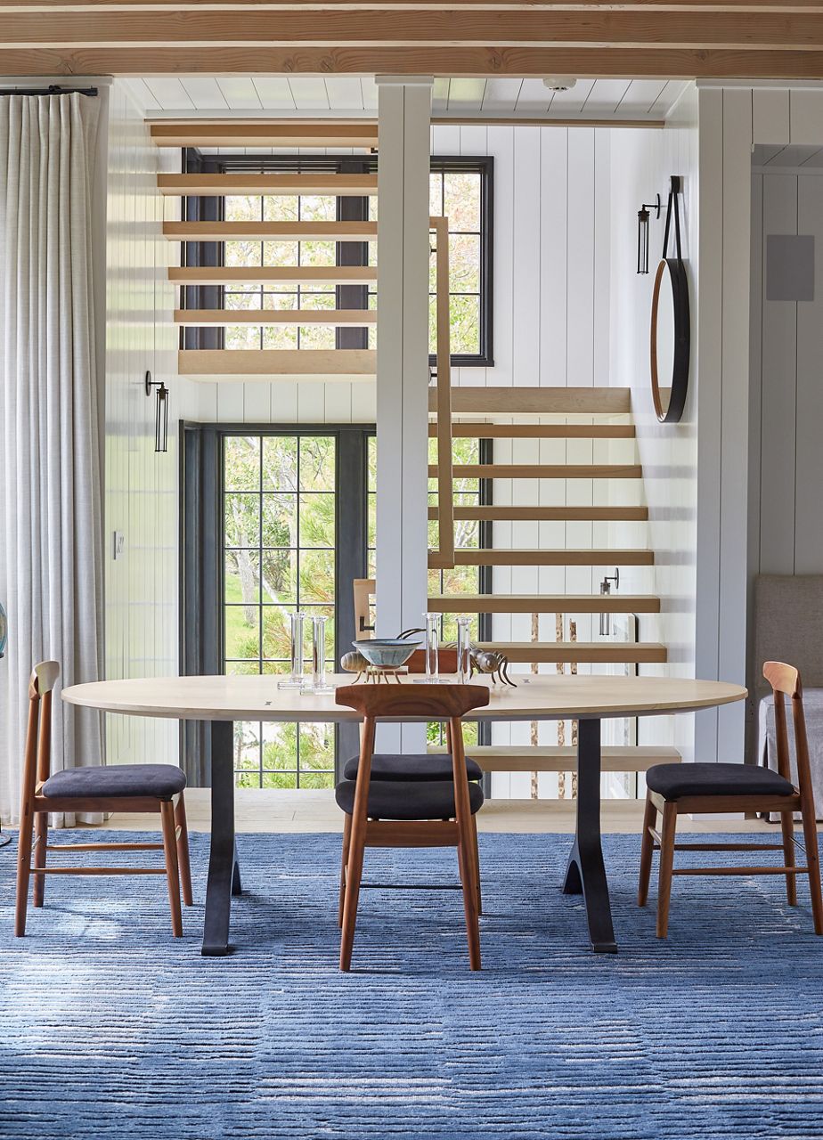 Une vue à travers une salle à manger avec un tapis bleu, des chaises en bois foncé avec des sièges bleus et une table ovale en bois pâle sur un escalier ouvert avec une lumière qui s’écoule à travers les fenêtres du plancher au plafond.