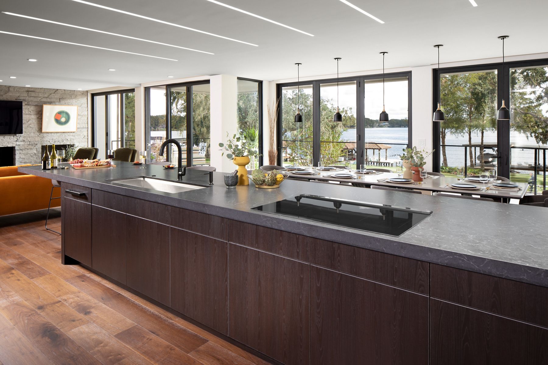 Daylight in large modern, open concept space featuring large kitchen island with cooktop, undermount sink, and Matte Black kitchen faucet with seating and dining areas in front of a wall of large windows overlooking lake.