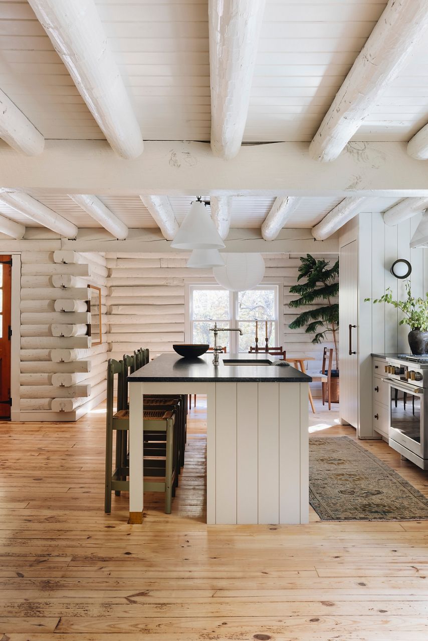 Wide image looking down the long way of the kitchen island, seeing the white wood base island seating area. The profile of the Kohler Artifacts faucet is centered on the island and between the sun-filled windows in the distance. Painted white logs of the cabin are visible on the walls and the ceiling. 
