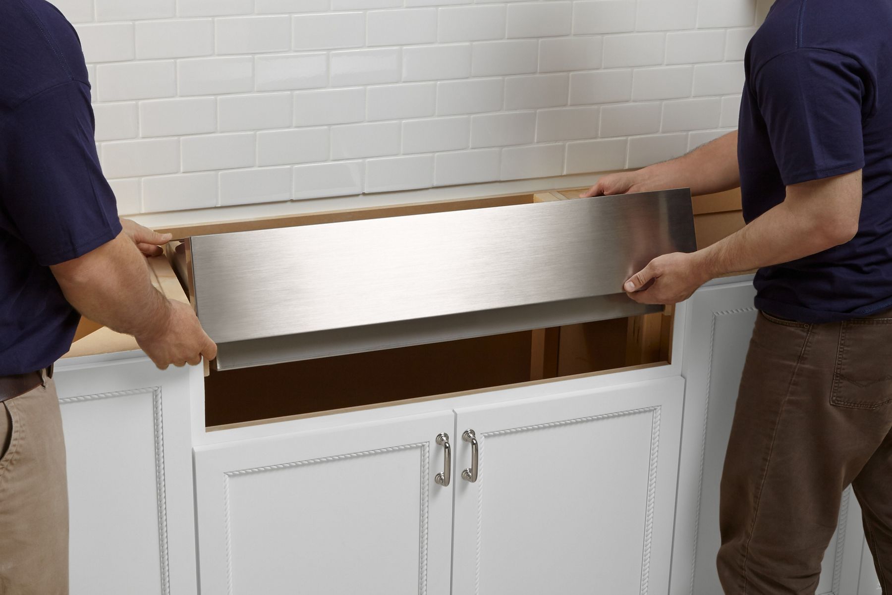 Two people install a KOHLER Vault stainless steel undermount kitchen sink with shortened apron-front into white cabinets with stainless steel drawer pulls