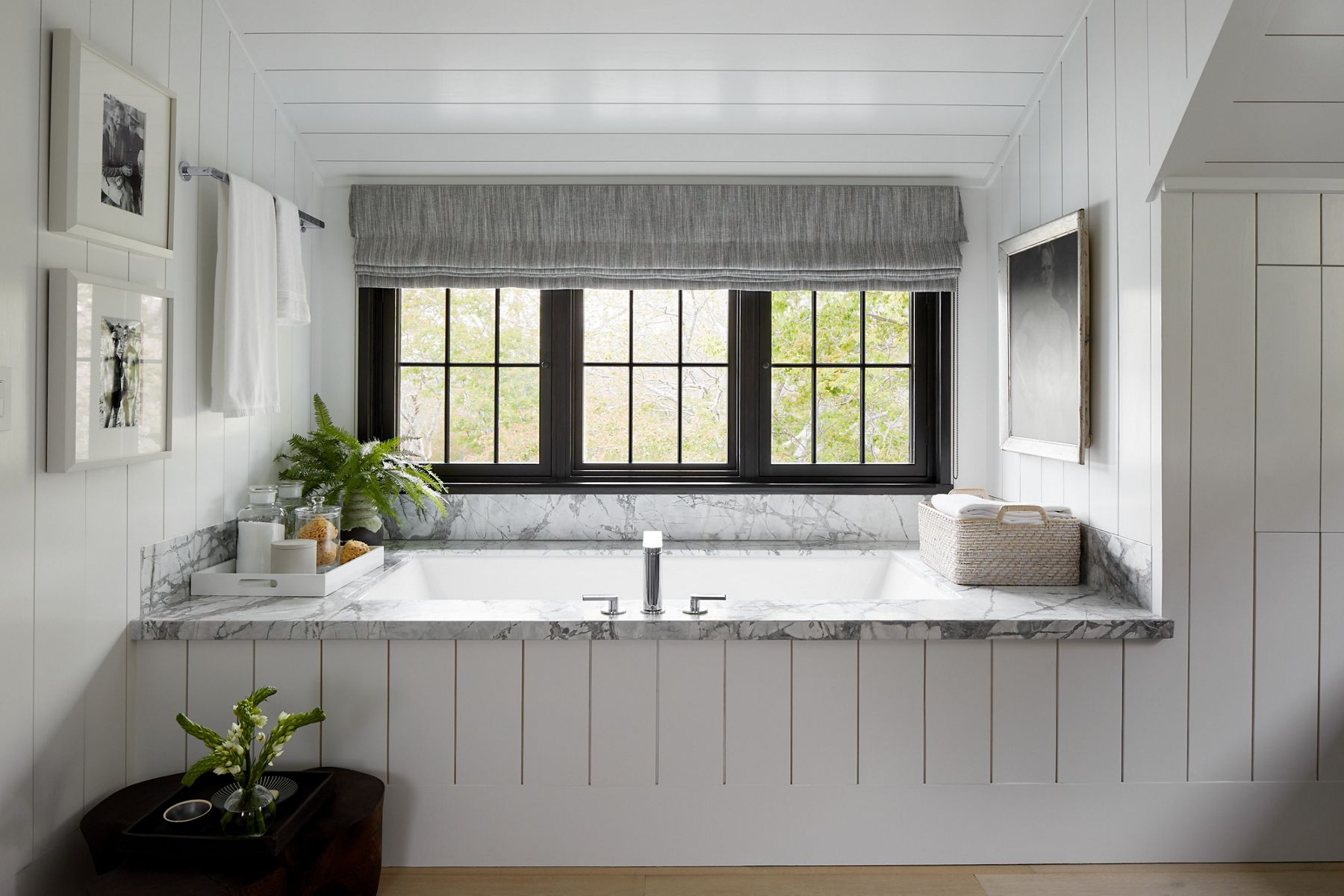 Une baignoire blanche en alcôve sous trois fenêtres lumineuses divisées à cadre noir, avec un contour de baignoire en planche blanche pour s’agencer aux murs blancs vides.