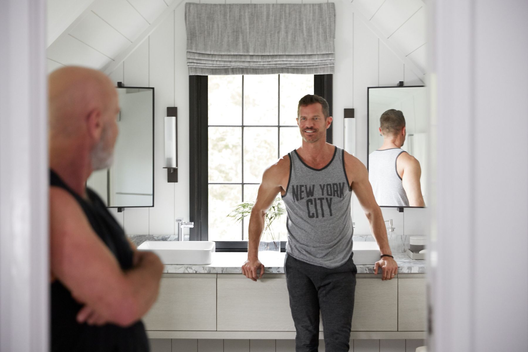 Ray et John se détendent dans une salle de bain dans un grenier avec des plafonds inclinés, une grande fenêtre centrale, un meuble-lavabo mural blanc avec 2 lavabos rectangulaires Vox et deux miroirs pivotants.