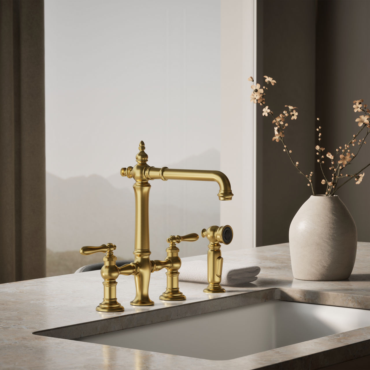 Polished gold kitchen bridge faucet with dual handles and side sprayer, mounted on a marble countertop beside a ceramic vase with branches.