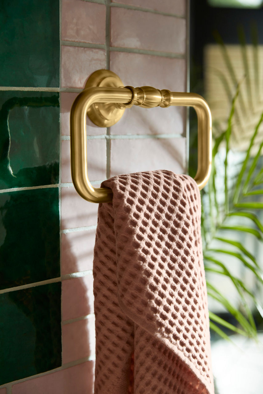 Polished gold towel ring with decorative detailing, holding a textured beige hand towel against green tile wall.