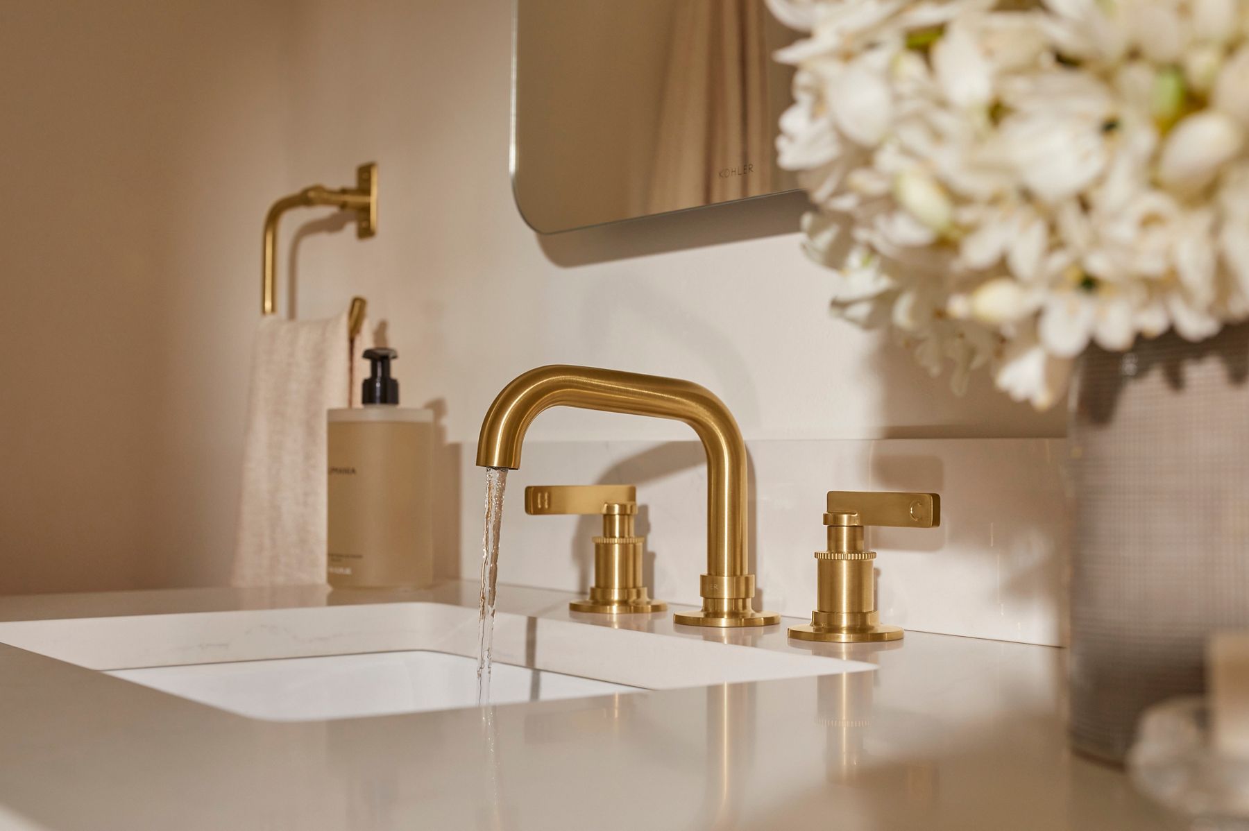 Castia collection brushed gold widespread bathroom faucet installed on a white vanity countertop with soft floral accents and a modern mirror in the background.