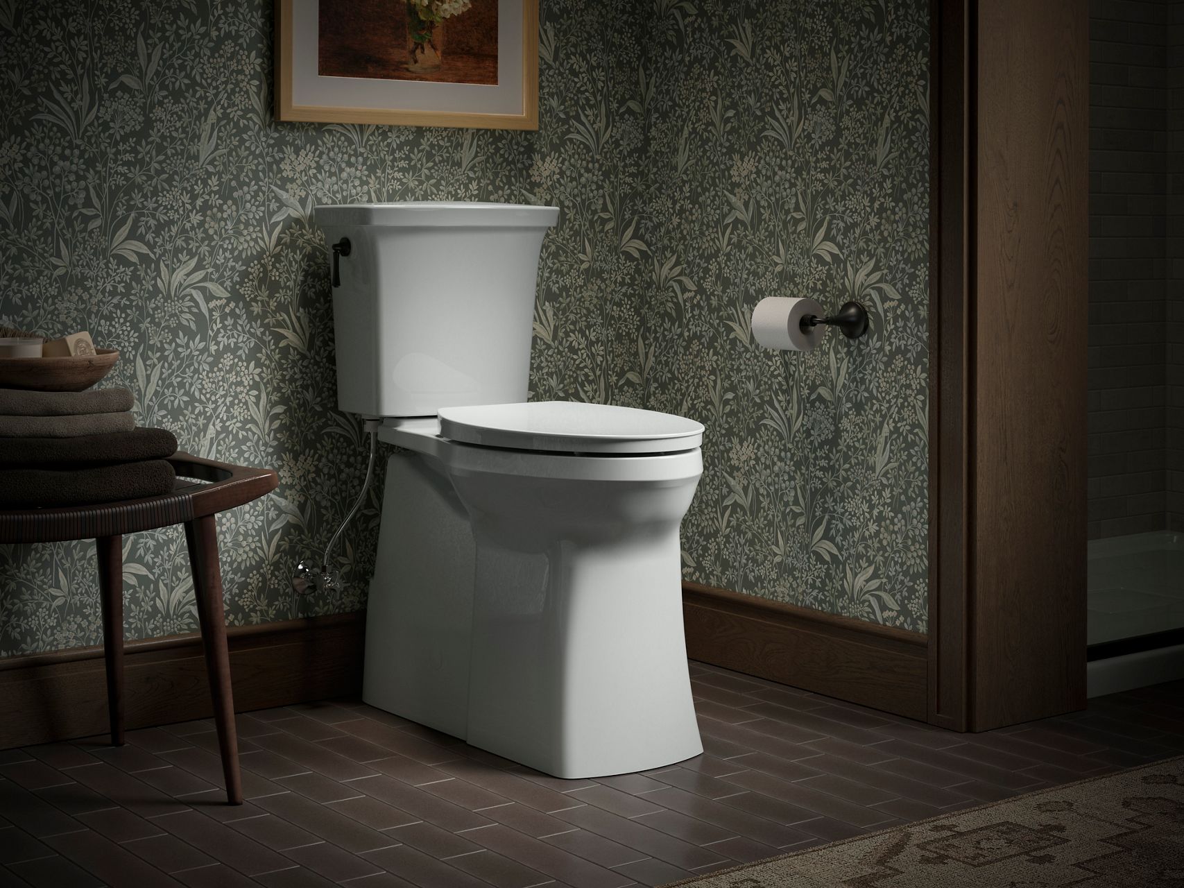 Traditional bathroom featuring a white toilet against patterned botanical wallpaper, with dark wood trim, tiled flooring, and a side table stacked with folded towels.