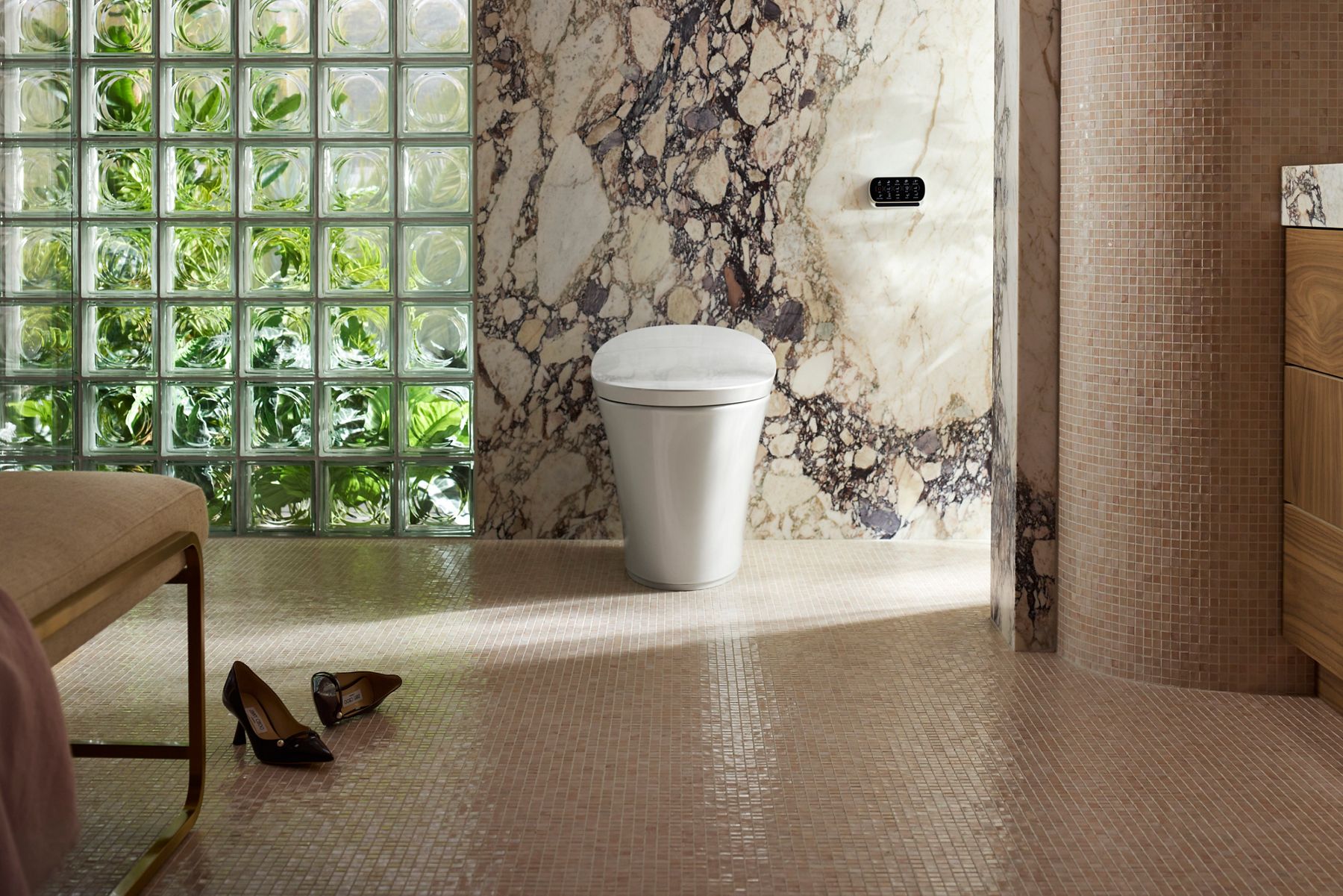 Modern bathroom featuring a white smart toilet set against a dramatic marble accent wall, with a glass block window, mosaic tile flooring, and warm natural light.