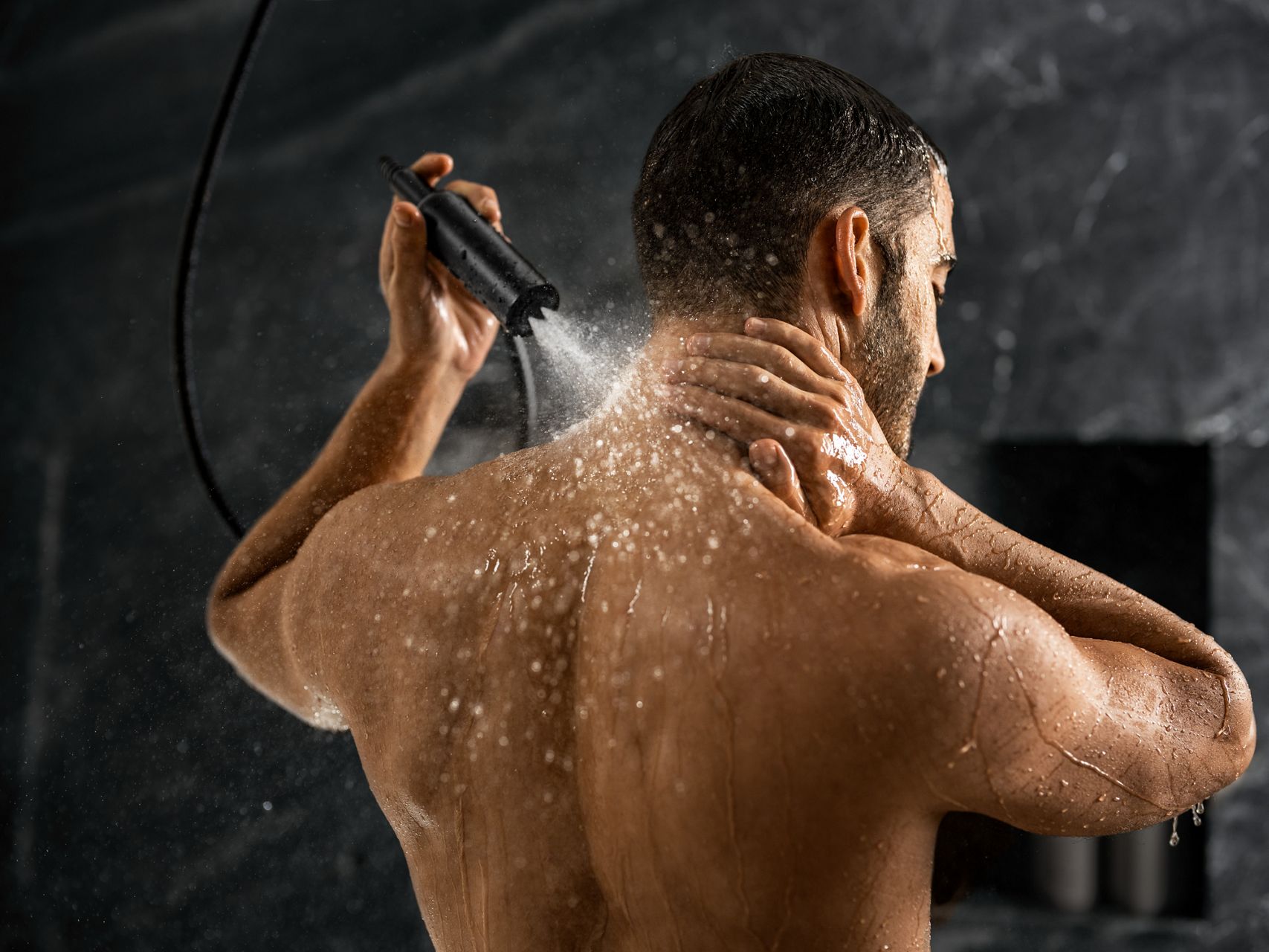 A man using a hand shower to spray water on the back of his neck.