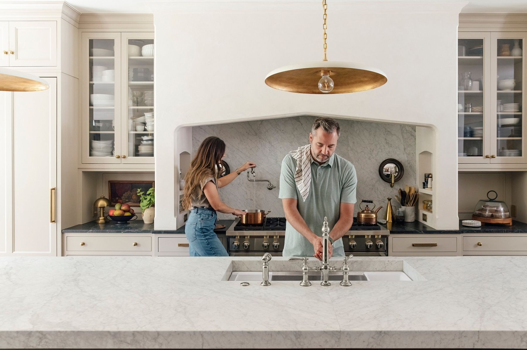 Wide image of Chris and Julia Marcum, the couple known online as ChrisLovesJulia, cooking in their large, open kitchen. Chris is standing behind the kitchen island sink with a towel over his shoulder and washing his hands in the workstation sink under the polished nickel Kohler Artifacts faucet, while Julia is standing at the stove behind him, filling a pot with water from the wall-mounted Kohler pot filler faucet.