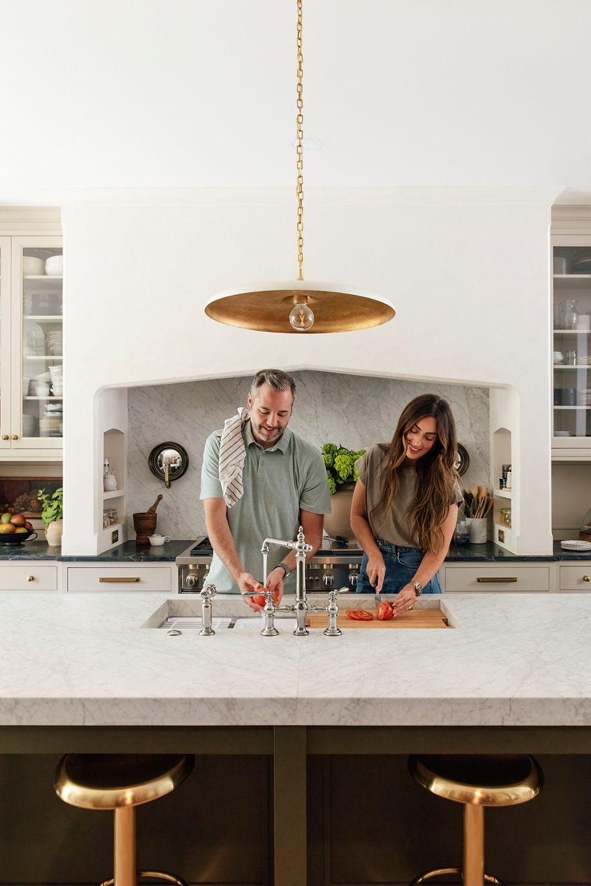Portrait of Chris and Julia Marcum standing in their kitchen, smiling and looking down at the food they are preparing in their kitchen. Chris is washing tomatoes under the Kohler kitchen sink faucet and Julia is cutting tomatoes on the cutting board accessory of the Kohler workstation kitchen sink.