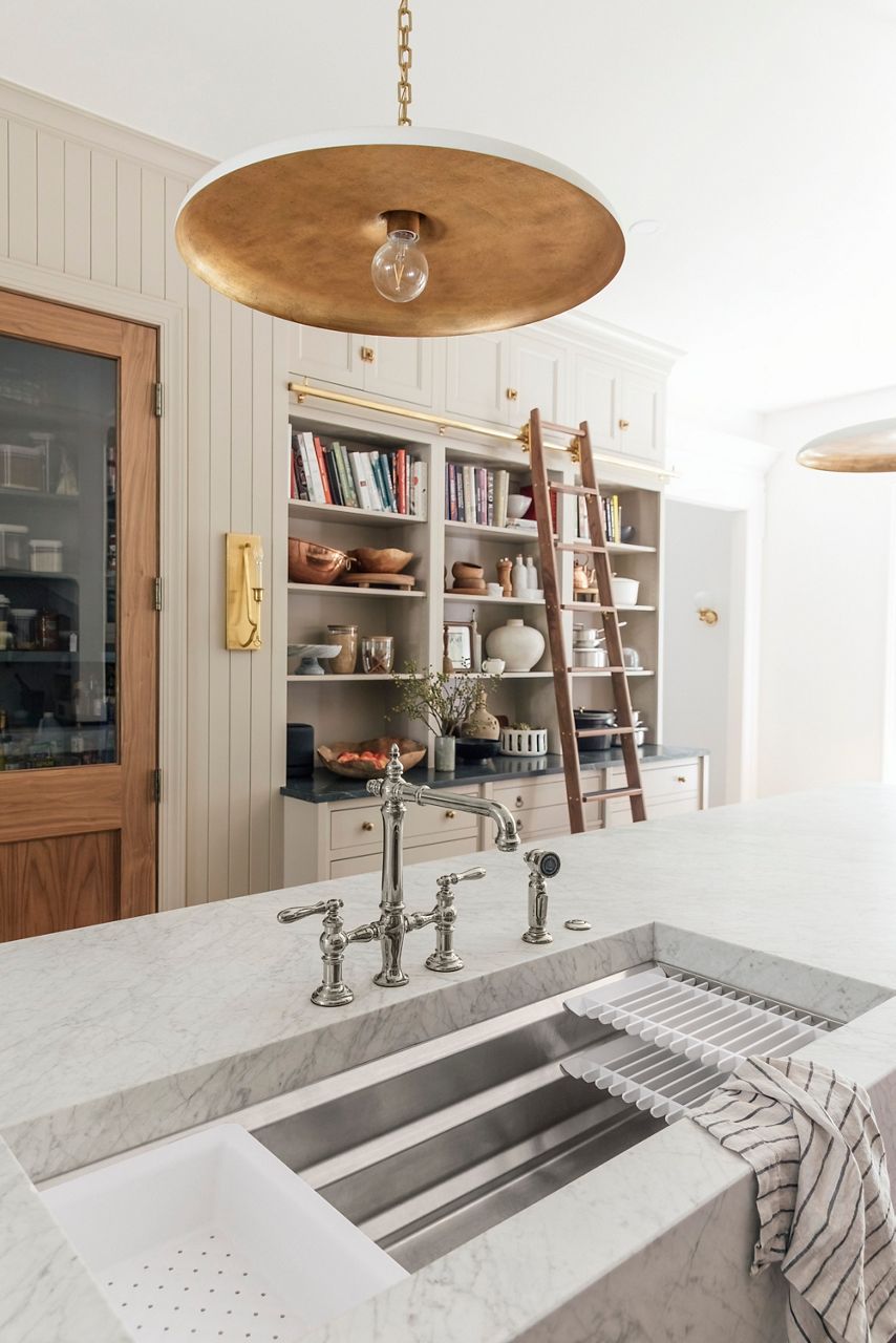 Image of Chris and Julia's kitchen, looking over the kitchen island sink featuring Kohler Artifacts faucet in polished nickel finish, where you can see the bookshelf and pantry door in the distance. The open shelves are filled with cookbooks, bowls and kitchen decor.