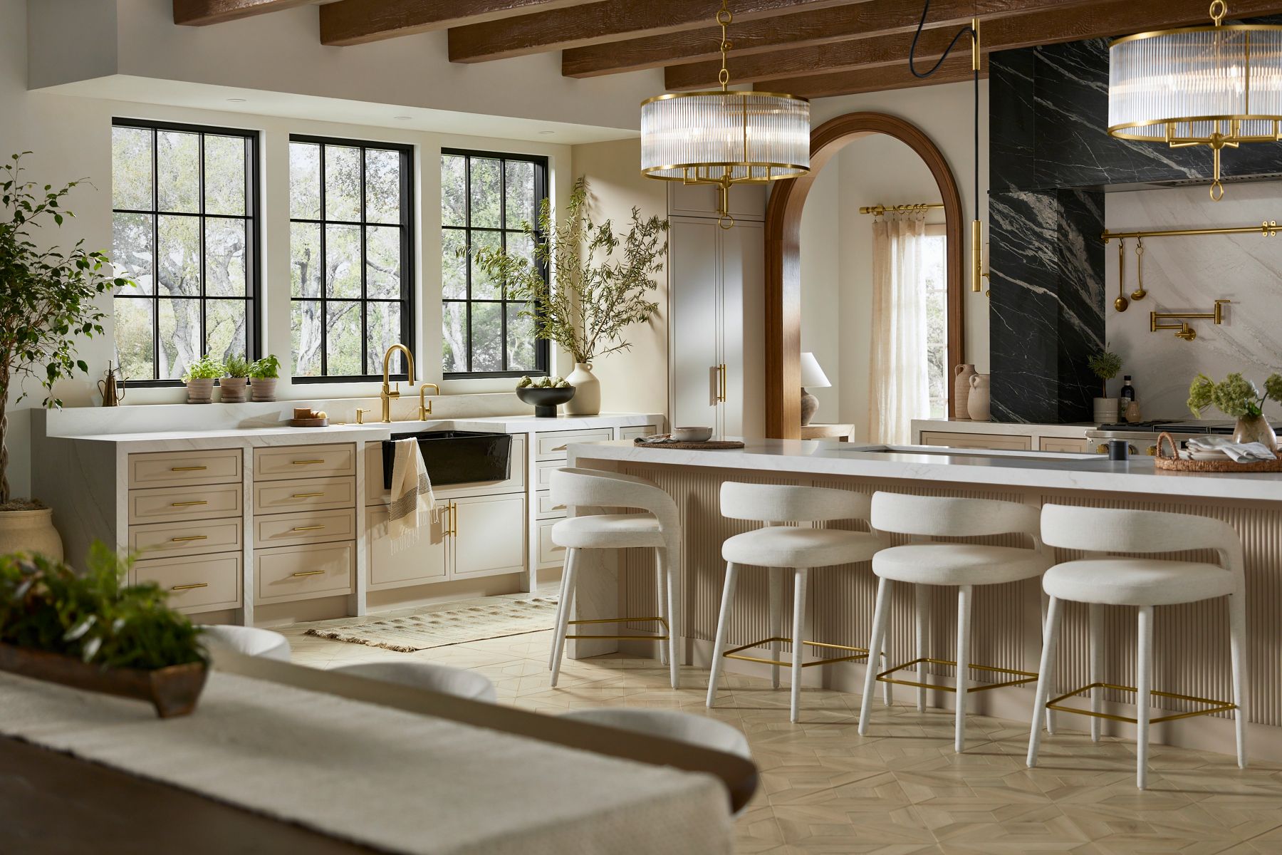 A large kitchen with an island in the center, a range top and oven behind it, a sink and cabinets on the left, and a refrigerator and wet bar on the right, all in neutral cream tones with gold and dark gray accents.