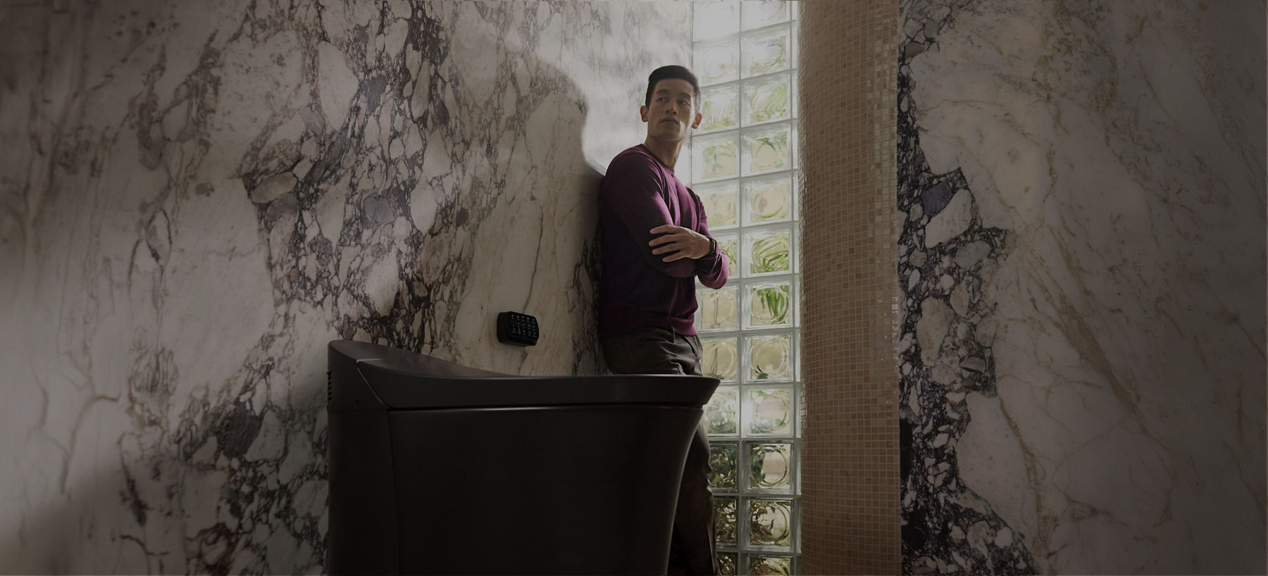 Modern space with marble walls and glass blocks; person in maroon sweater stands near a KOHLER Smart Toilet.