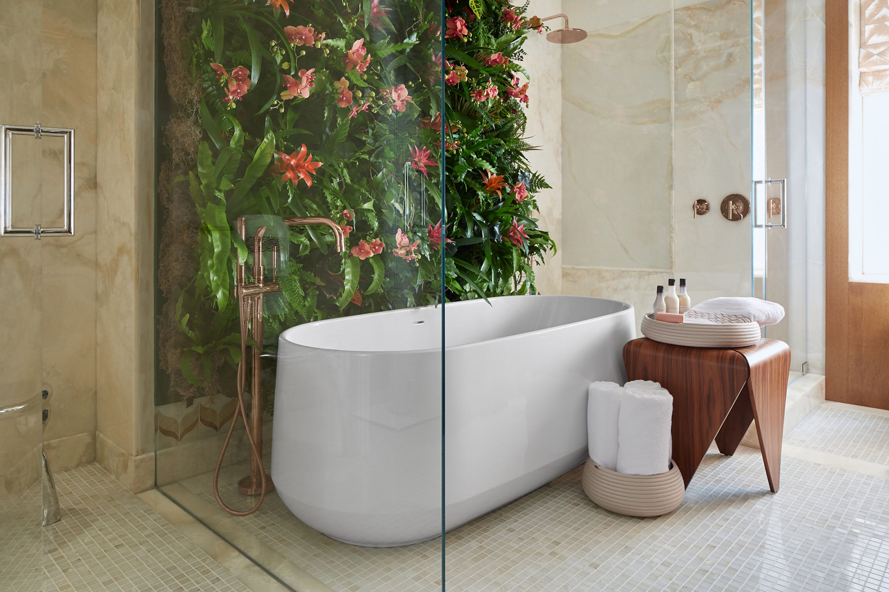 A cream-colored bathroom with a white freestanding bath with a wall of plant life on its left and a wood table with bathing supplies to its right.
